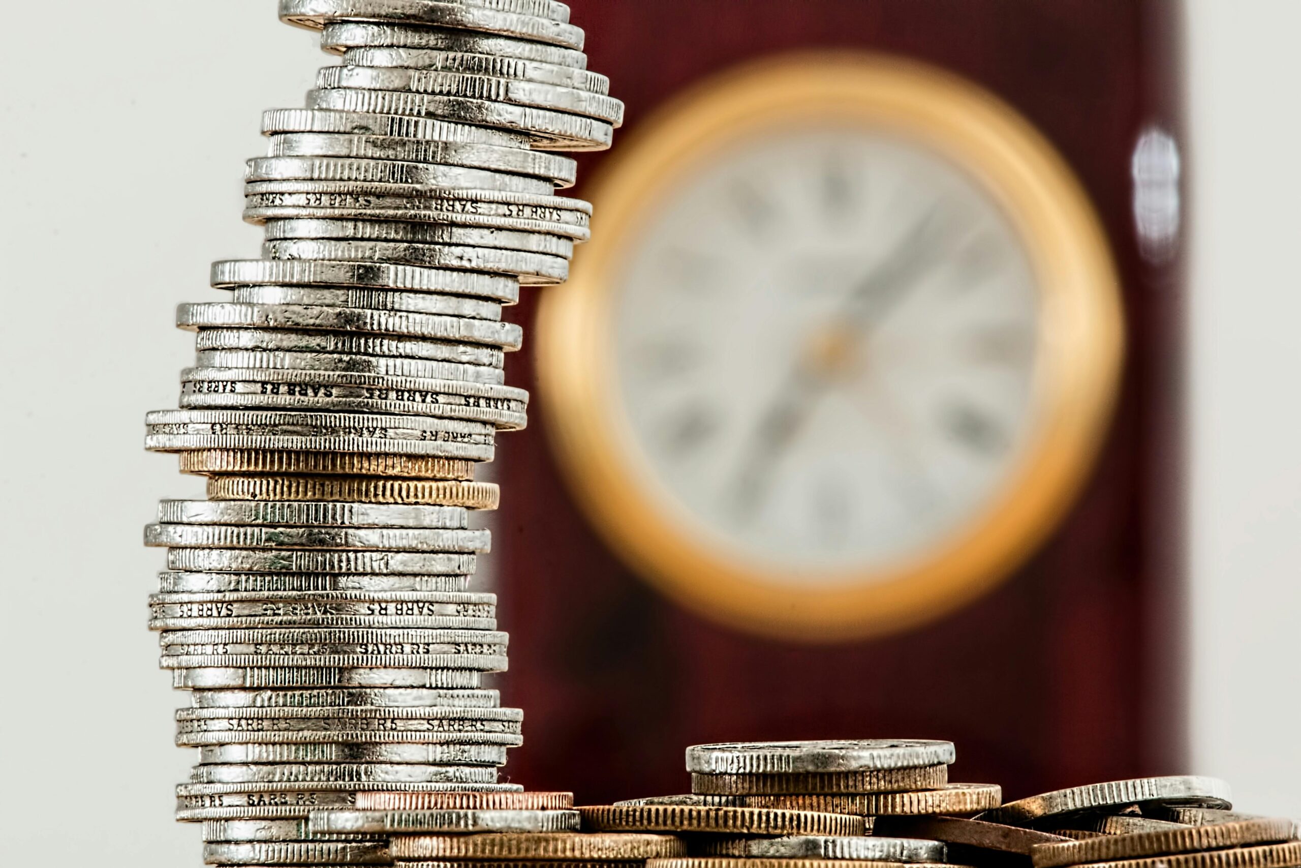 Home A close-up image of stacked coins with a blurred clock, symbolizing time and money relationship.